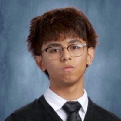 Portrait of a young man wearing glasses, a black blazer, and black tie against a blue background