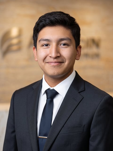 Professional headshot of an Asian man in a dark business suit, white dress shirt, and dark tie, smiling at the camera against a blurred beige background
