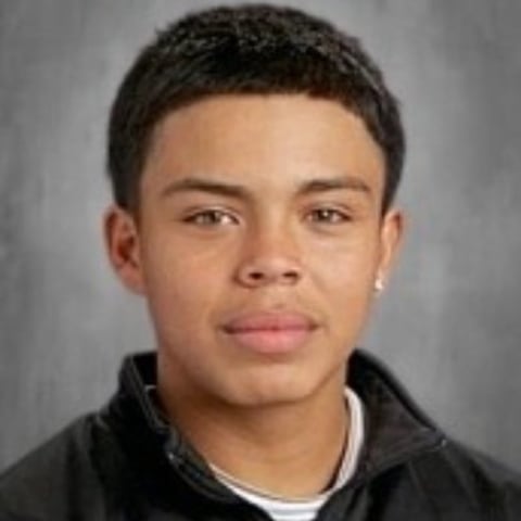Portrait of a young man with short dark hair wearing a black jacket over a white shirt against a gray background